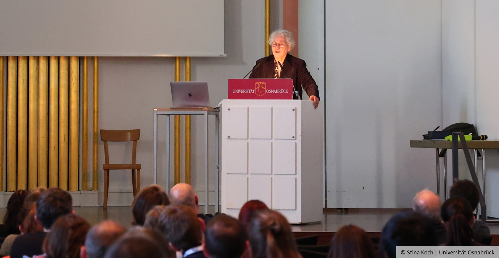 Prof. Dr. Nüsslein-Volhard stands at a lectern and speaks, in the foreground you can see part of her audience from behind.