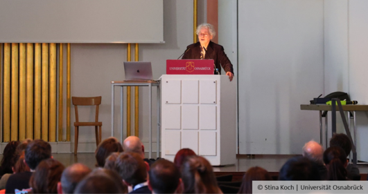 Prof. Dr. Nüsslein-Volhard stands at a lectern and speaks, in the foreground you can see part of her audience from behind.