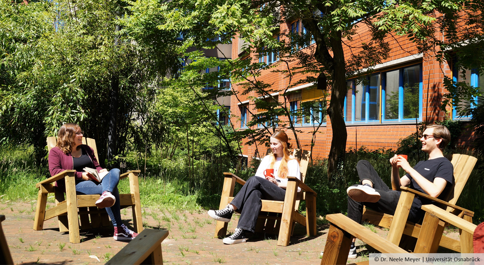 Studierende sitzen auf Hölzmöbeln im begrünten Innenhof und unterhalten sich.