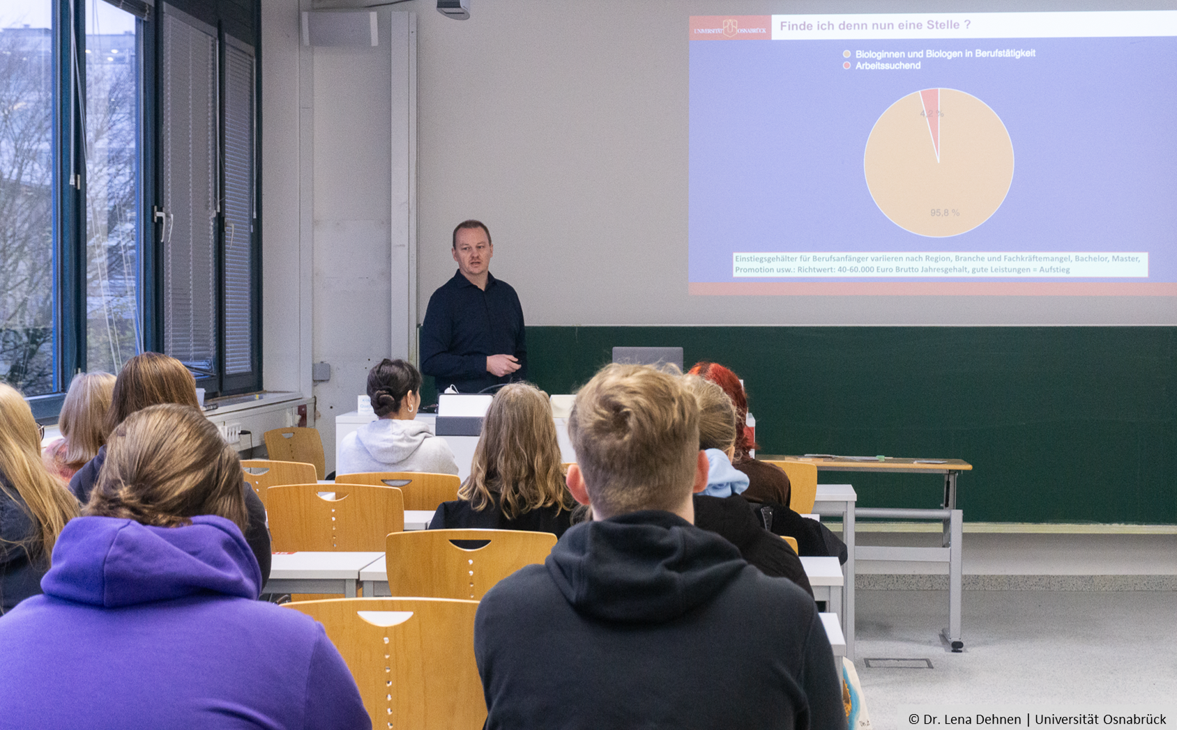 A man is standing in front of pupils sitting on chairs and is giving a lecture.