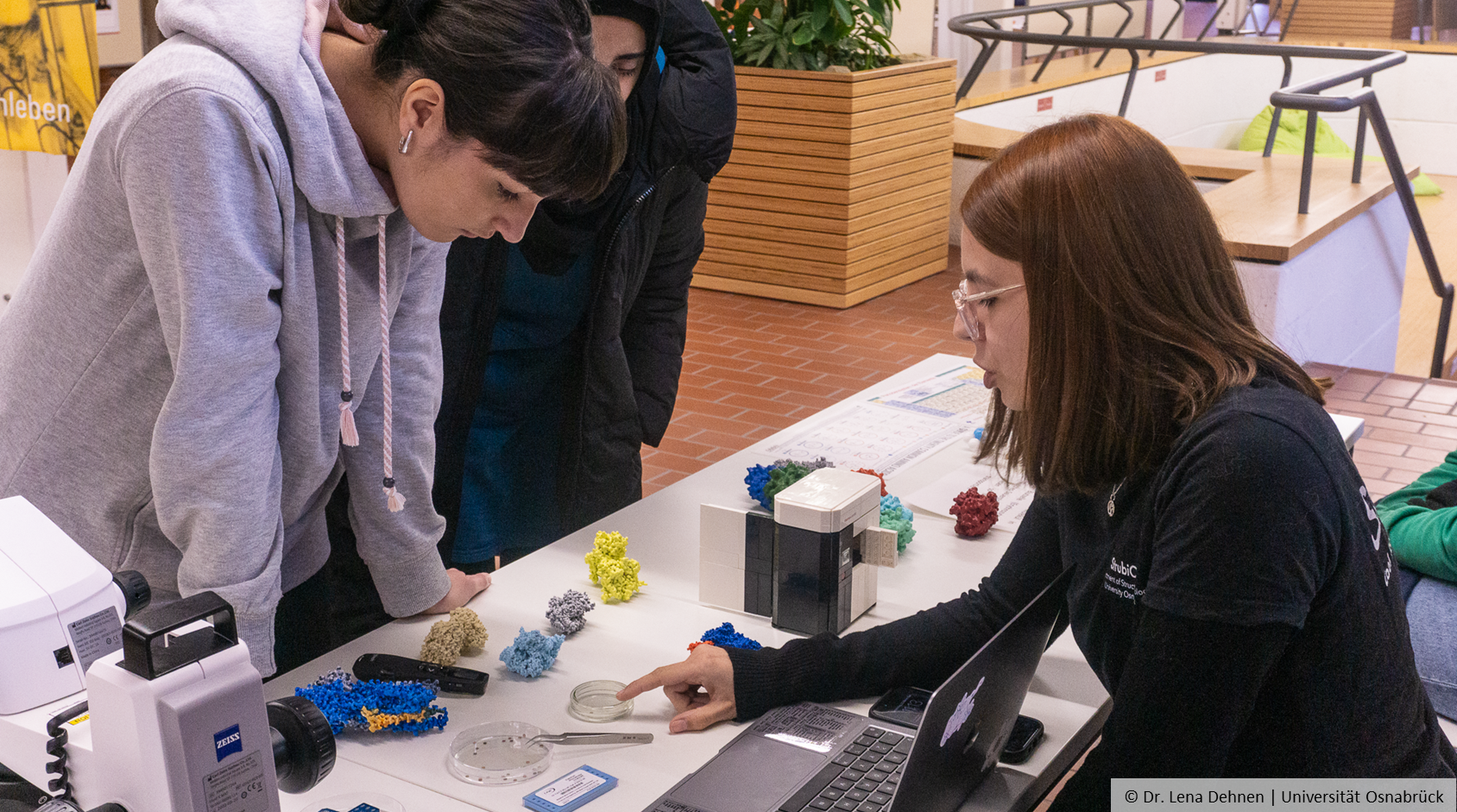 There are models of molecules on a table with pupils standing in front of them. A person on the other side points to a small bowl.