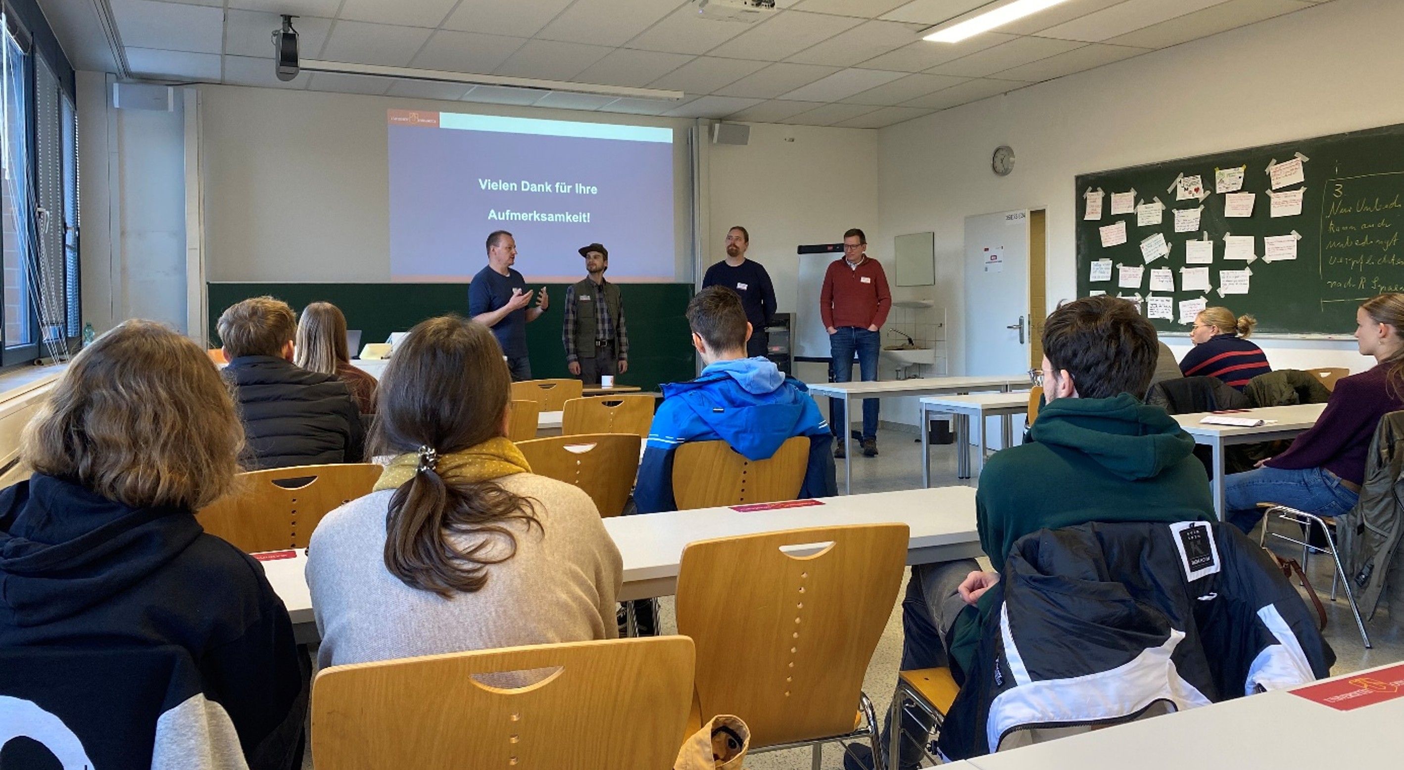 Four men are standing in a seminar room where pupils are sitting