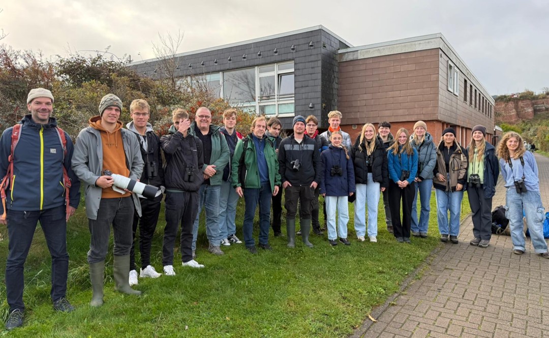 Around twenty people are standing in front of a building, some carrying binoculars.