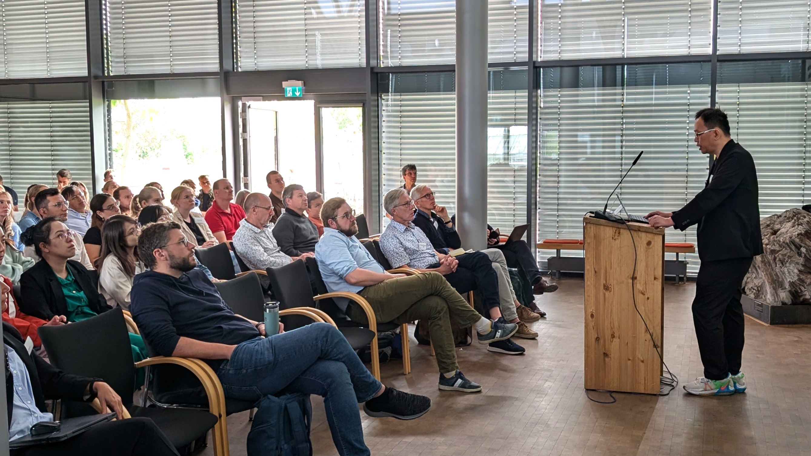 A speaker stands at a lectern, with people sitting on chairs in front of him.