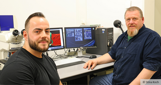 Zwei Männer sitzen vor einem Labortisch. Im Hintergrund sind ein Mikroskop und Bildschirme zu sehen.