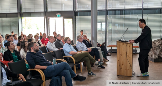 A speaker stands at a lectern, with people sitting on chairs in front of him.