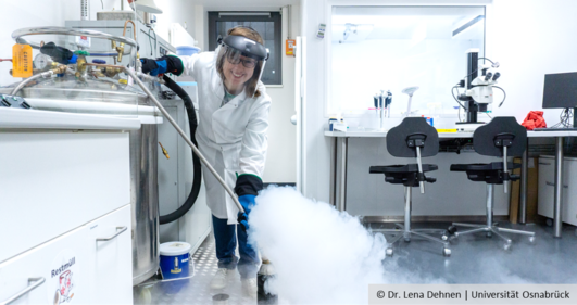 A woman stands in a laboratory wearing a lab coat, safety goggles and thick gloves, with steam rising in front of her.