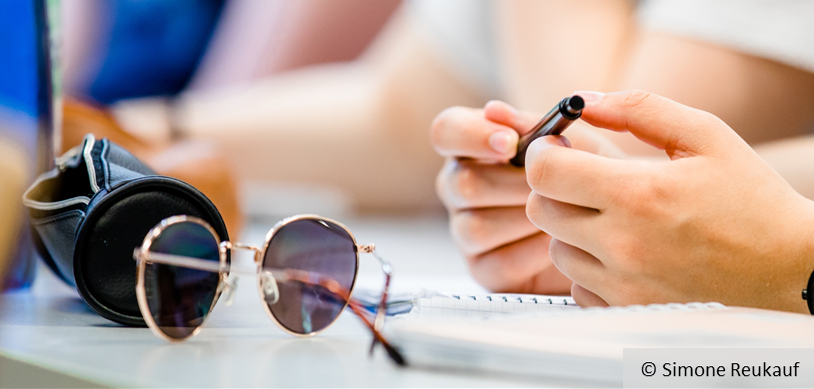 Ein Etui, eine Sonnenbrille und eine Hand, die einen Stift hält, © Simone Reukauf